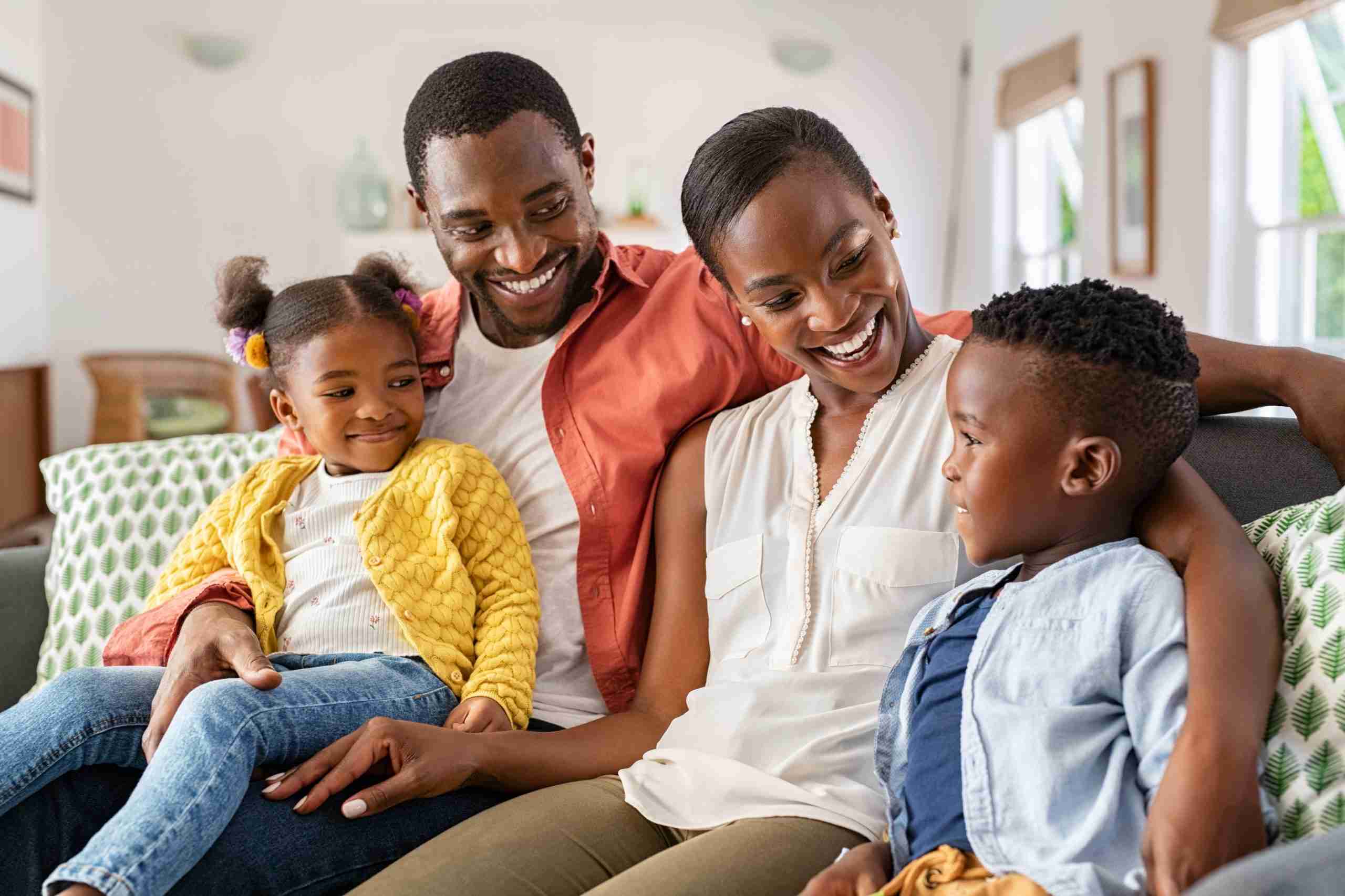 Smiling African-American family relaxing on couch and talking to each other. Happy black father and mother sitting on sofa at home with their daughter and son. Playful ethnic family with two little kids sit on couch cuddling.