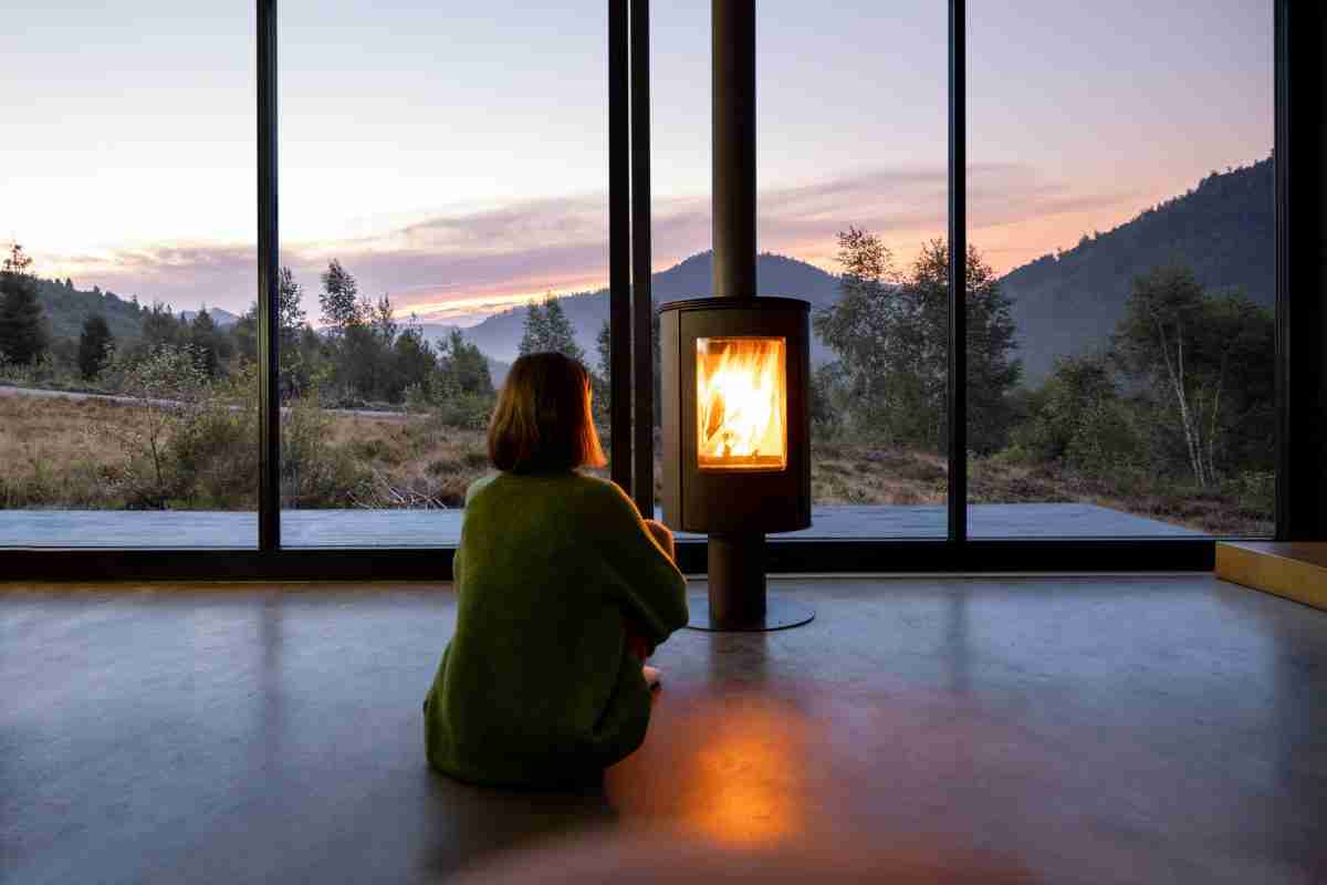 Woman enjoy great view on mountains while sitting near fireplace at modern living room at sunset . Concept of rest in houses or cabins on nature. Solitude in nature and escape from everyday life